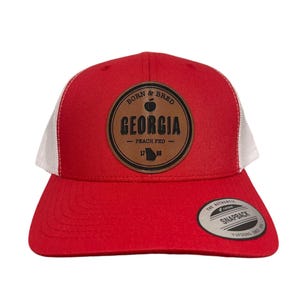 May include: Red and white trucker hat with a brown leather patch that says "Born & Bred Georgia Peach Fed 1788" with an apple and the outline of the state of Georgia.