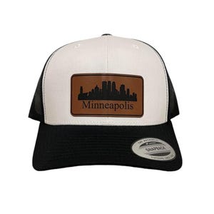 Op de afbeelding: Een witte en zwarte truckerpet met een bruin leren patch met de skyline van Minneapolis en de naam van de stad. De pet heeft een zwarte mesh achterkant en een gebogen klep. De Snapback-sluiting is zichtbaar.