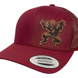Puede incluir: Gorra de camionero de color rojo burdeos con un parche de cuero marrón que presenta un monstruo de dibujos animados con una amplia sonrisa. La gorra tiene una parte trasera de malla y un cierre a presión. La etiqueta de la gorra dice "The Authentic Yupoong Snapback Yupoong Since 1974."