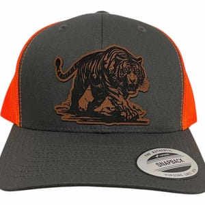 Op de afbeelding: Grijze en oranje truckerpet met een leren patch met een gedetailleerd tijgerontwerp. De pet heeft een gebogen klep, een mesh achterkant en een snapbacksluiting. De patch toont een tijger die loopt.