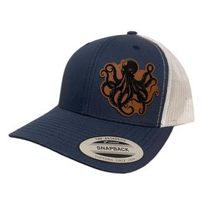 May include: A navy blue and white trucker hat with a brown leather patch featuring a black octopus design. The hat has a snap closure and the text "The Authentic Yupoong Snapback Yupoong Since 1974" on the back.
