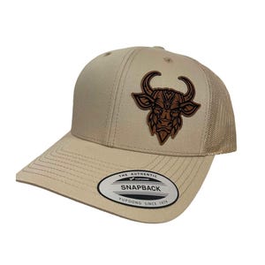 May include: A tan and brown trucker hat with a brown leather bull head patch. The hat has a mesh back and a snap closure. The text "THE AUTHENTIC YUPOONG SNAPBACK YUPOONG SINCE 1974" is printed on the hat.