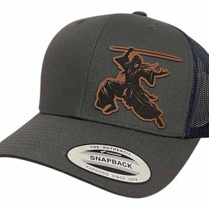 May include: A charcoal gray Snapback baseball cap with a dark blue mesh back. The cap features a brown leather patch with a black silhouette of a samurai warrior. The text "THE AUTHENTIC SNAPBACK YUPOONG SINCE 1974" is on the front.