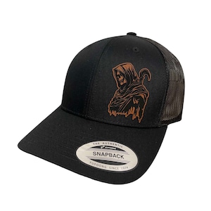 May include: Black and brown trucker hat with a brown leather patch featuring a hooded figure with a scythe. The hat has a black mesh back and a black snap closure. The patch reads "The Authentic Snapback Yupoong Since 1974".