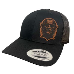 Może przedstawiać: Czarna i brązowa czapka trucker z naszywką skórzaną przedstawiającą vintage'ową ilustrację wampira. Czapka ma czarny siateczkowy tył i regulowane zapięcie na zatrzask. Czapka ma białą metkę z tekstem "THE AUTHENTIC YUPOONG SNAPBACK YUPOONG SINCE 1974".