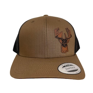 May include: A brown and black trucker hat with a brown leather deer head patch. The hat has a black mesh back and a brown brim. The hat has a "The Authentic Snapback" label on the back.