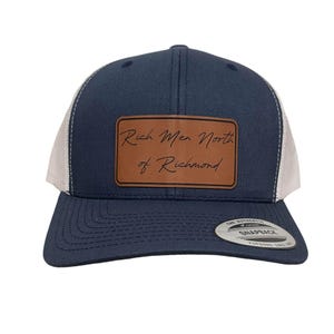 Puede incluir: Una gorra de camionero azul marino y blanca con un parche de cuero marrón que dice "Rich Men North of Richmond".