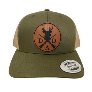 May include: Olive green and tan trucker hat with a leather patch featuring a deer silhouette, crossed rifles, and the letters "DAD." The hat has a curved brim and a snapback closure. A white and silver tag is attached to the brim.