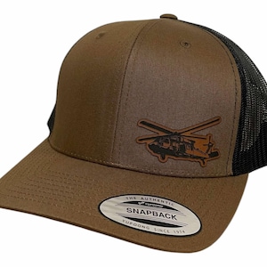 May include: A brown and black trucker hat with a black mesh back. The hat features a brown leather patch with a black silhouette of a helicopter on it. The hat also has a white label on the bottom that says "The Authentic Yupoong Snapback Since 1974."