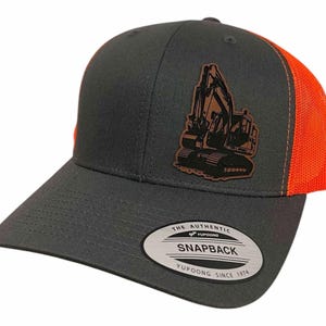 May include: A gray and orange trucker hat with a brown leather patch featuring a silhouette of an excavator. The hat has a mesh back and a snap closure. The patch has the text "THE AUTHENTIC" and "YUPOONG" on it. The hat also has a white label with the text "THE AUTHENTIC YUPOONG SNAPBACK YUPOONG SINCE 1974" on it.