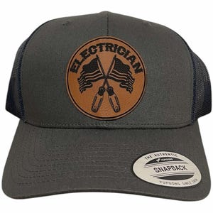 May include: A gray and black trucker hat with a brown leather patch that says "Electrician" with two crossed screwdrivers and American flags. The hat has a black mesh back and a Yupoong logo.