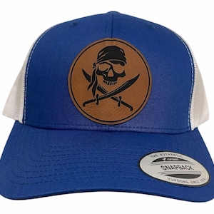 May include: A royal blue trucker hat with a white mesh back. The hat features a brown leather patch with a black skull and crossbones design. The patch has the text "The Authentic Snapback Yupoong Since 1974".