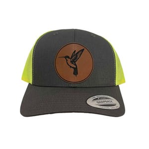 Puede incluir: Una gorra de camionero gris oscuro con una malla amarilla en la parte posterior. La gorra tiene un parche de cuero marrón con un diseño de colibrí negro.