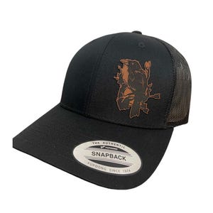 May include: Black and brown trucker hat with a leather patch featuring a bird on a branch. The hat has a black mesh back and a black snap closure. The hat has a label that reads "The Authentic Yupoong Snapback Yupoong Since 1974".