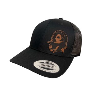 Puede incluir: Gorra de camionero negra y marrón con un parche de cuero que presenta la cara de una mujer. La gorra tiene una parte trasera de malla negra y un cierre de presión negro. La gorra tiene el texto "THE AUTHENTIC YUPOONG SNAPBACK YUPOONG SINCE 1974" en la parte posterior.