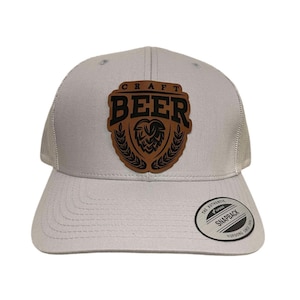 May include: A gray and brown baseball cap with a leather patch that says "CRAFT BEER" with a hop graphic. The cap is a snapback style.