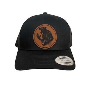 May include: Black trucker hat with a brown leather patch featuring a werewolf design. The hat has a curved brim and a snapback closure. The patch shows a detailed illustration of a snarling werewolf.