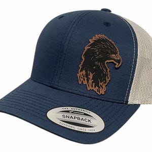 Op de afbeelding: Een navyblauwe en witte truckercap met een bruine leren adelaarspatch. De pet heeft een mesh achterkant en een gebogen klep. De pet heeft een label met de tekst "The Authentic Yupoong Snapback Yupoong Since 1974".