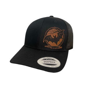 May include: Black and brown baseball cap with a leather patch featuring a bat silhouette in front of a full moon. The patch says "The Authentic Snapback Yupoong Since 1978".