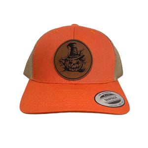 May include: Orange and brown trucker hat with a leather patch featuring a black and white illustration of a pumpkin wearing a witch's hat. The patch is round and has a stitched edge. The hat has a mesh back and a snap closure.