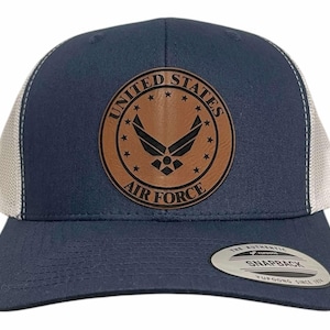 May include: A navy blue and white trucker hat with a brown leather patch displaying the United States Air Force emblem. The patch has the words "UNITED STATES AIR FORCE" and stars. The hat has a snapback closure.