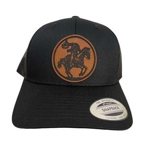 May include: Black trucker hat with a brown leather patch featuring a silhouette of a person riding a horse and holding a jack-o-lantern. The patch is circular and has a brown background. The hat has a black mesh back and a black brim. The hat has a white label on the back that says "The Authentic Yupoong Snapback Yupoong Since 1974."