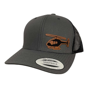May include: A charcoal gray Snapback baseball cap with a black mesh back and a curved bill. The cap features a brown leather patch with a black helicopter silhouette. The tag reads "THE AUTHENTIC SNAPBACK YUPOONG SINCE 1974."