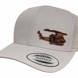 May include: A light gray snapback hat with a brown helicopter patch. The hat has a mesh back and a curved bill. The patch features a detailed illustration of a helicopter. The hat has the text "The Authentic Snapback Yupoong Since 1974".