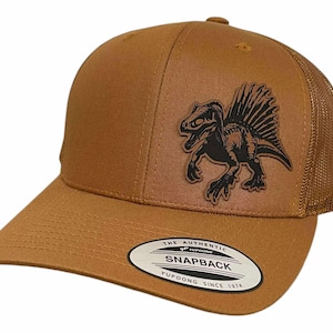 May include: A brown Snapback baseball cap with a mesh back and a curved peak. The cap features a black dinosaur graphic on the front and the text "THE AUTHENTIC SNAPBACK YUPOONG SINCE 1974" on the peak.