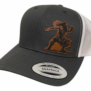 Könnte beinhalten: Dunkelgraue Snapback-Kappe mit weißem Mesh-Rücken und einem braunen Lederaufnäher mit einer indianischen Figur. Die Kappe hat einen gebogenen Schirm und einen Schnappverschluss.