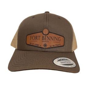 Op de afbeelding: Een bruine en beige trucker-pet met een bruin leren patch waarop staat "FORT BENNING GEORGIA 32° 21' 8.5297° N 84° 58' 7.7407° W".