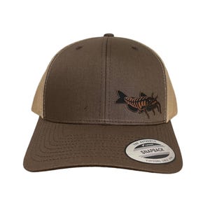 Könnte beinhalten: Eine braune und beige Trucker-Kappe mit einem braunen Wels-Motiv und dem Text "The Authentic Yupoong Snapback Yupoong Since 1974" auf der Rückseite.