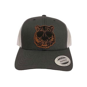 Op de afbeelding: Een donkergrijze en witte truckercap met een bruine leren patch van een tijger met een zonnebril. De pet heeft een wit mesh-achterpaneel en de tekst "The Authentic Snapback" op de achterkant.