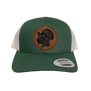 May include: A green and white trucker hat with a brown leather patch featuring a black silhouette of a turkey. The patch has the text "The Authentic Snapback Yupoong Since 1974".