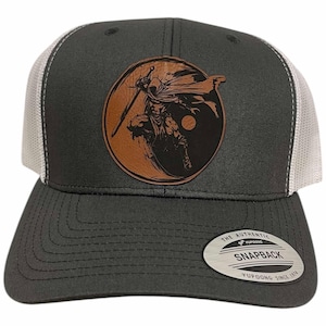 Peut inclure: Un chapeau de camionneur gris et blanc avec un patch en cuir marron représentant une illustration en noir et blanc d'une figure tenant une épée. Le patch est circulaire et a un symbole yin yang en arrière-plan. Le chapeau a un dos en maille et une fermeture à pression. Le texte "THE AUTHENTIC SNAPBACK YUPOONG SINCE 1974" est imprimé sur une étiquette argentée attachée au chapeau.