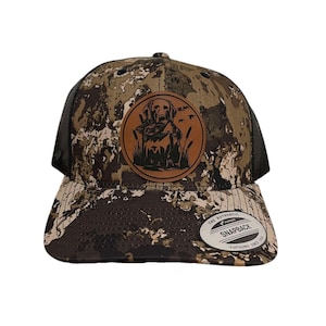 Puede incluir: Una gorra de camionero de camuflaje marrón y negro con un parche de cuero marrón que presenta una silueta negra de un perro con un pato en la boca. El parche está rodeado de un círculo verde con juncos marrones. La gorra tiene una parte trasera de malla negra y una correa ajustable marrón con el texto "THE AUTHENTIC SNAPBACK" en blanco.