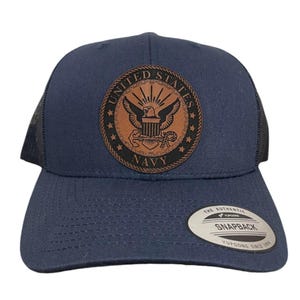 Puede incluir: Una gorra de camionero azul marino con un parche de cuero marrón que presenta el sello de la Marina de los Estados Unidos. El parche tiene las palabras "United States Navy" alrededor del sello.