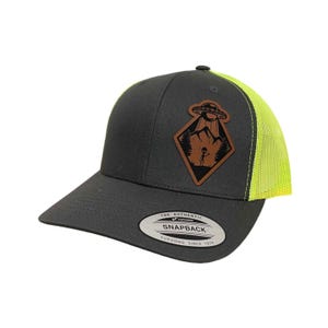 Puede incluir: Una gorra de camionero gris y amarillo neón con un parche de cuero marrón que presenta un OVNI, montañas y una figura de palo. El parche tiene forma de diamante y tiene el texto "THE AUTHENTIC SNAPBACK YUPOONG SINCE 1974".