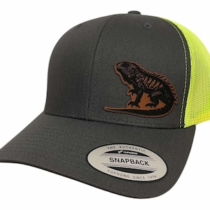May include: A grey and neon yellow trucker hat with a brown leather iguana patch. The hat has a mesh back and a snap closure. The hat has a label that reads "The Authentic Yupoong Snapback Yupoong Since 1974".