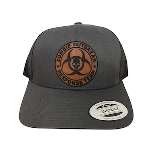 May include: A gray and black trucker hat with a brown leather patch that says "Zombie Outbreak Response Team" with a skull and biohazard symbol. The hat has a black mesh back and a "Yupoong The Authentic Snapback" label.