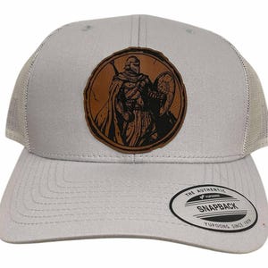 May include: A light gray trucker hat with a mesh back and a curved brim. The front features a circular brown leather patch with a detailed illustration of a warrior holding a shield and sword. The hat has a snapback closure.