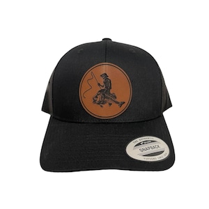 May include: Black trucker hat with a brown leather patch featuring a silhouette of a person fishing. The patch has the text "THE AUTHENTIC SNAPBACK YUPOONG SINCE 1974".
