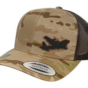 Op de afbeelding: Een beige en bruine camouflage trucker cap met een zwarte vliegtuigpatch en een bruin label "The Authentic Snapback Yupoong Since 1974".