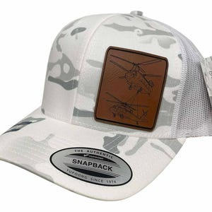 Op de afbeelding: Witte en grijze camouflage Snapback pet met een bruin leren patch met helikopter illustraties. De pet heeft een witte mesh achterkant en een gebogen klep. Het label luidt "MULTICAM".