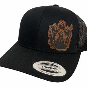 May include: Black trucker hat with a mesh back and a curved brim. The front features a brown leather patch with a graphic of several grim reapers. The hat has a snapback closure and a Yupoong tag.