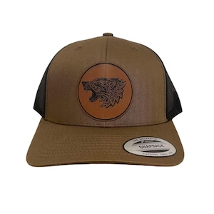 May include: A brown and black trucker hat with a brown leather patch featuring a black and white wolf design. The patch is circular and has the text "The Authentic Snapback" on the back of the hat.