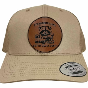 Puede incluir: Gorra de camionero color beige con parte trasera de malla y visera curva. La parte delantera presenta un parche de cuero marrón con un gráfico de mapache y el texto "Strawberry Jams But My Glock Don't". Una etiqueta Snapback está en la visera.