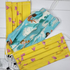May include: Three pleated face masks with adjustable ear loops. Two masks are yellow with pink bird patterns. The third mask is blue with a surfing scene. All masks are made of fabric.