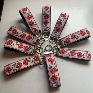 May include: A selection of key fobs with a floral pattern. Each fob has a white strap decorated with red flowers and green leaves. The key fobs have a metal ring and a black backing.