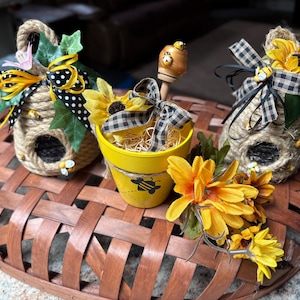 May include: Decorative bee-themed items on a brown woven basket. Includes two rope beehive ornaments with sunflowers and bows, a yellow pot with a bee graphic, and a wooden honey dipper. Artificial flowers and greenery add color.